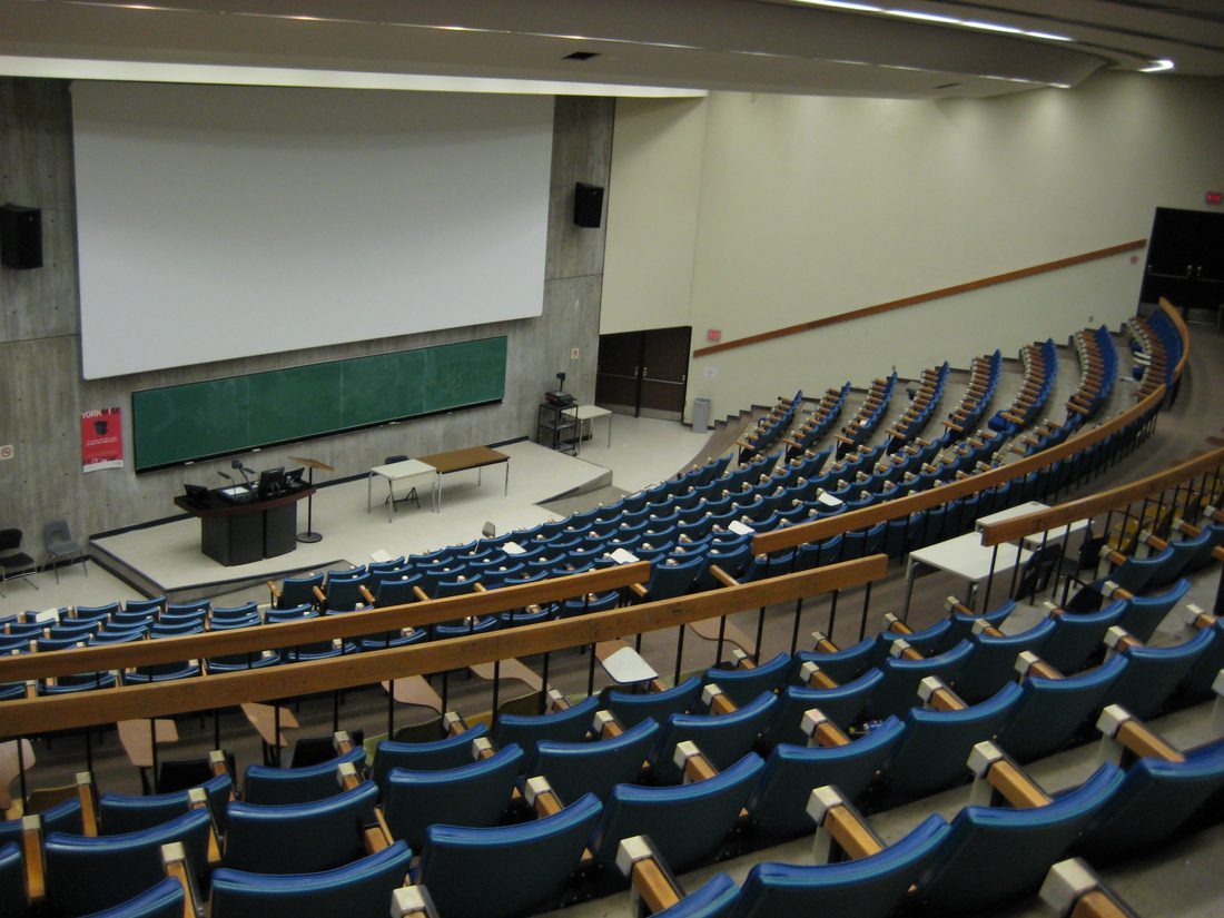 An empty university lecture hall with tiered blue seating facing a lectern and projection screen, illustrating the classroom format international students must adapt to.